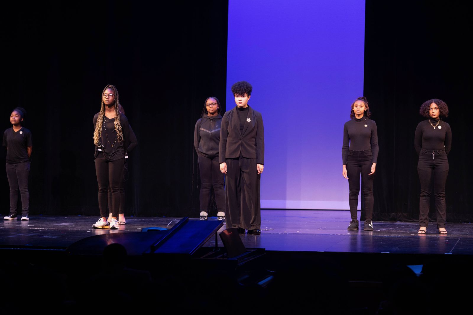 Young performers dressed in black stand in still rows on a stage lit by a violet backdrop, with a grand piano at the stage edge.