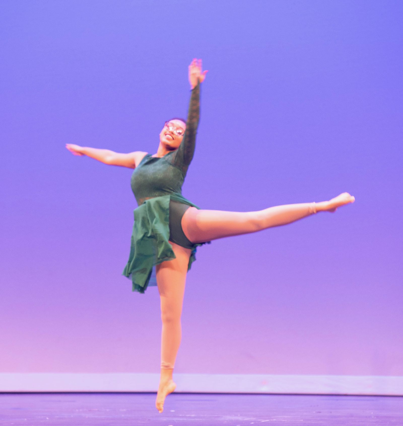A smiling dancer in a green costume leaps with one leg extended and an arm raised against a violet backdrop.