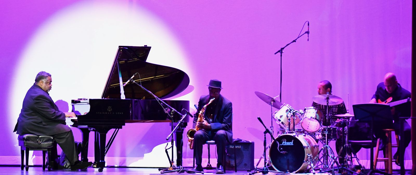 Alvin Waddles leads a quartet of grand piano, saxophone, drums, and guitar in a single spotlight on a magenta-lit stage.